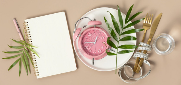粉红色的盘子装有餐具板测量胶带空白纸笔记本水热带叶和按颜色背景平地顶视图复制空间的闹钟背景