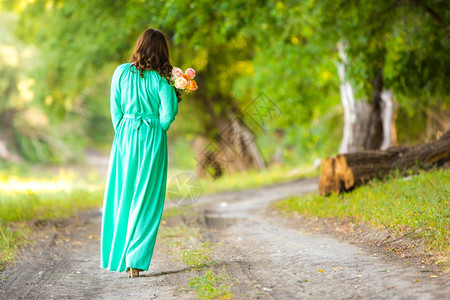 在一个温暖的夏日树林里走在一个穿着长裙盛一束花的女孩在路上走到了远处图片