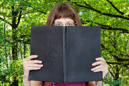 带着眼镜的女孩看着大书上面有空白的封绿树林在背景上图片