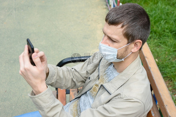 一个戴着保护医疗面具的男子坐在公园长凳上用手机进行交流图片