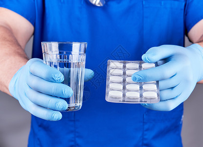 穿蓝制服和不消毒的乳胶手套医生持有一杯水和包白药丸图片