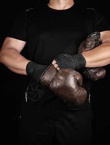 黑色衣服的成年肌肉男在比赛前双手戴上棕色皮衣拳击手套双被裹在黑色运动绷带中图片