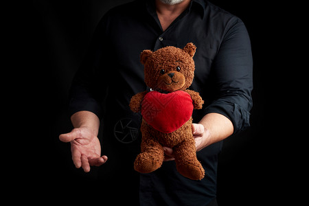 身着黑色衬衫的成年男子在深色背景上抱着一只红心的棕色泰迪熊低调地祝贺2月14日情人节的到来图片