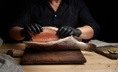 黑衬衫主厨和乳胶手套在棕色木制切板上挂着一具无头鲑鱼的原始,这是切鱼的过程。图片