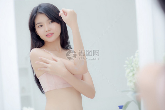 美丽感的年轻女看着镜子的美丽感年轻女将奶油涂在手臂和信息上美貌女孩运用身体润肤皮护理健康软水合液治疗和化妆品与润湿剂图片