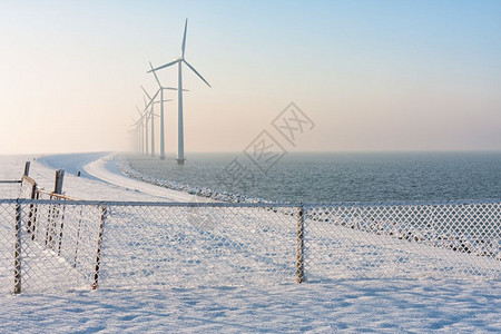 在冬季荷兰在河中与雪栅栏和长排风力涡轮机在冬季烟雾中缺水的离岸停靠长排风力涡轮机一起冬荷兰在季用雪围栏和风力涡轮机起水图片