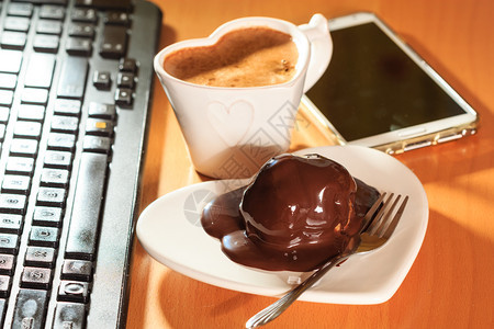 在笔记本电脑键盘和智能手机旁边的心形板碟子上咖啡和巧克力蛋糕办公室工作台图片