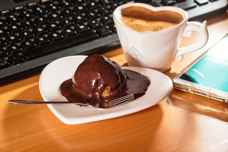 在笔记本电脑键盘和智能手机旁边的心形板碟子上咖啡和巧克力蛋糕办公室工作台图片