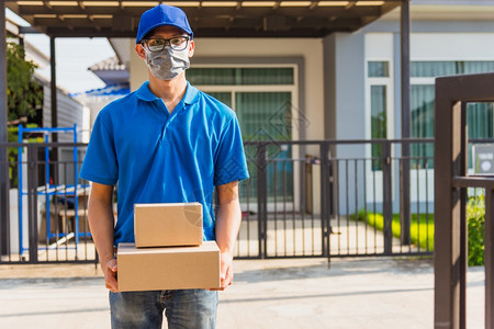 亚洲青年送货员在线亚洲青年送货员箱装蓝帽子和T恤衫他保护面罩根据宵禁检疫大流行冠状COVID19服务客户在房子门前图片