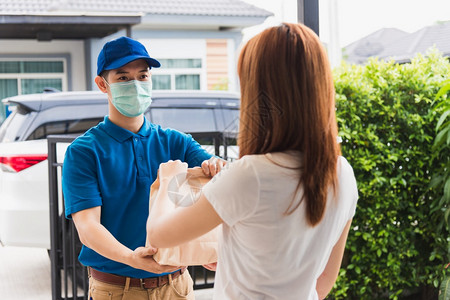 亚洲快送递的年轻男子向接受保护面罩的女客户提供纸袋快餐根据宵禁检疫大流行冠状COVID19进行保护面罩图片