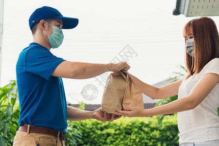 亚洲快送递的年轻男子向接受保护面罩的女客户提供纸袋快餐根据宵禁检疫大流行冠状COVID19进行保护面罩图片