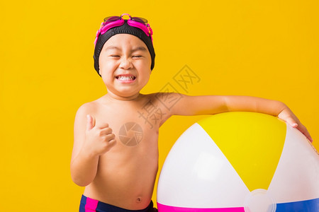暑假概念肖写亚洲快乐可爱的小孩男笑在泳衣里握着海滩球举起手指拇以示好兆头摄影棚拍孤立的黄色背景图片