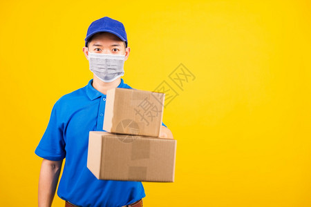 身穿蓝色T恤衫和制服的亚洲年轻送货工人前线身穿蓝色T恤衫和帽子制服的亚洲年轻送货工人身穿面罩保护在冠状或COVID19下用纸板盒背景图片