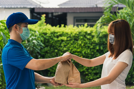 亚洲快送递的年轻男子向接受保护面罩的女客户提供纸袋快餐根据宵禁检疫大流行冠状COVID19进行保护面罩图片