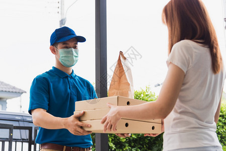 亚洲快送的青年男子递向接受保护面罩的女客户提供纸袋快餐和披萨盒根据宵禁检疫大流行冠状COVID19图片