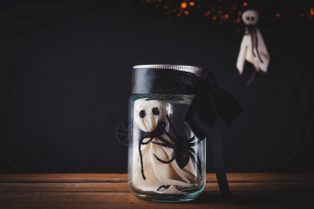 有趣的万圣节装饰晚会白色幽灵恐怖脸庞和黑蜘蛛在木制桌上的罐子玻璃杯中摄影棚拍孤立在黑色背景上快乐节的概念图片