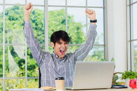 很高兴奋的亚洲男子举起手臂庆祝成功年轻的商人在办公桌使用笔记本电脑很高兴从工作中获得丰厚的赢家利润图片