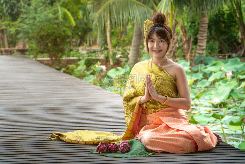 一种身着传统泰式礼服坐在木桥上的女美丽她手在泰国风格下举手受人尊敬在莲花叶上还有粉红莲美丽的传统图片