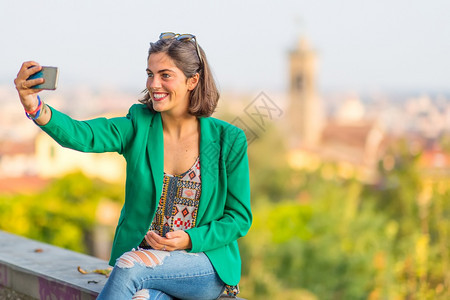 城市女移动的带着手机自拍年轻美丽笑女孩与本市背景人物一起使用手机图片