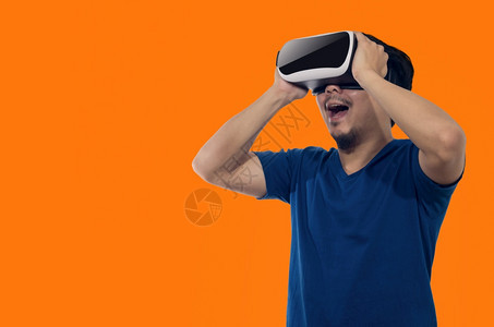 游戏玩家兴奋的白色有虚拟现实头盔的年轻人站在橙色背景的室内青年拿着虚拟现实头盔的年轻人图片