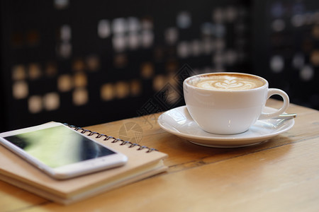 咖啡店用智能手机在木桌上配有拿铁艺术的白色咖啡杯食物卡布奇诺香气图片