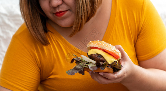 饥饿超重的年轻亚洲女人拿着汉堡包她饿了所有的时间和过量吃饱谷食和狂她的生活方式正在吃快餐所有时间不健康的概念美国人烧烤面包背景图片