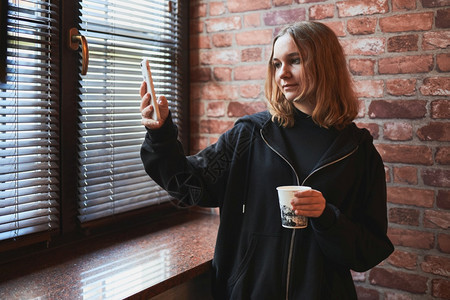 聊天在线的年轻女有视频电话远程交谈自拍照持智能手机的自拍喝咖啡放松同时在办公室休息站在窗户边上发言的女孩联系背景图片