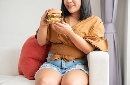 请享用饥饿的超重妇女笑着拿汉堡包坐在客厅里她非常快乐并享受吃餐狂食饮紊乱概念一顿饭重量图片