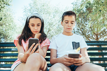 儿童坐在公园的长椅上看手机他们着技术玩人们图片