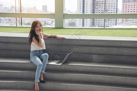 女孩学习白衬衫蓝色牛仔衣黑长头发使用电话和脑笔记本坐在楼梯上工作场所图片