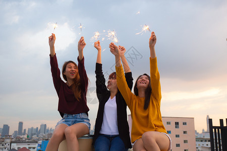 高兴地亚裔朋友点燃火花日落时享受自由年轻女在屋顶上快乐地举着火花保持户外图片