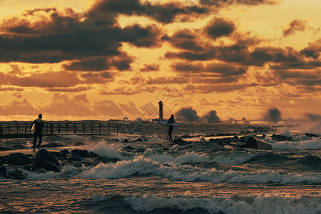 跑步有两个无法辨认的风筝冲浪者在暴风海码头周围奔跑日落光照耀着黄昏的阳光在戏剧橙色夜云下幕后有灯塔的轮影美丽面目全非图片