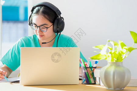 快乐的女士带着眼镜耳机的亚洲女学生少笑着看书本笔记电脑上的写笔记在桌子上从家中大学班级习在线研究教育亚裔妇女在书本网上学习时写笔图片