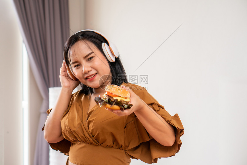 使用耳机的饥饿超重妇女笑着拿汉堡包坐在客厅里她非常快乐并享受吃餐狂饮食紊乱BED概念胖的房间可口图片