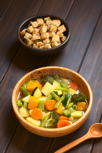 由青绿豆胡萝卜椰菜花土豆和南瓜制成的木碗蔬菜汤背面有一小碗包条在黑木上用天然灯光拍攝自面包丁健康图片