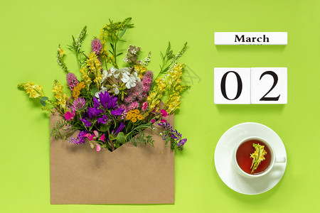 提醒躺3月2日Wooden立方体日历2杯草药茶Kraft信封里装着绿色背景的多彩花朵你好春天创意之顶风光平板木林立方体日历3月2图片