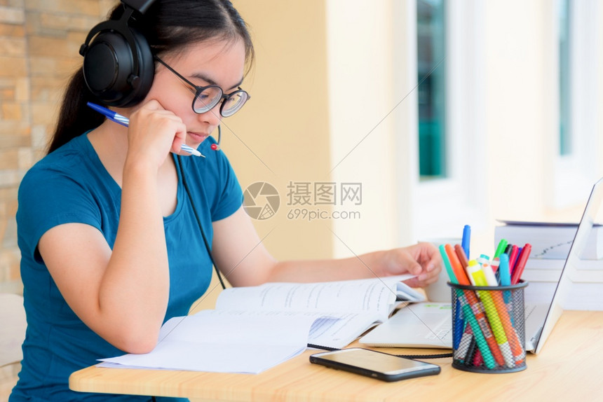 亚洲女学生少带着眼镜耳机坐看认真阅读一本书的烦恼使用笔记本电脑在桌子上学习的笔记本电脑进行在线学习从家中大班级的亚裔妇女认真阅读图片