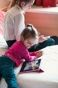 肖像药片白种人少女使用手机与妹一起在平板上观看动电影这两个女孩都坐在卧室的床上她们同的妹一起在平板电脑上看动画影图片