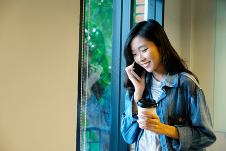 移动的亚洲人快乐女在握着咖啡杯站在窗口边聊天时欢乐的亚裔办公室女孩在讲手机时微笑人们在电话通讯技术生活方式概念等常设图片