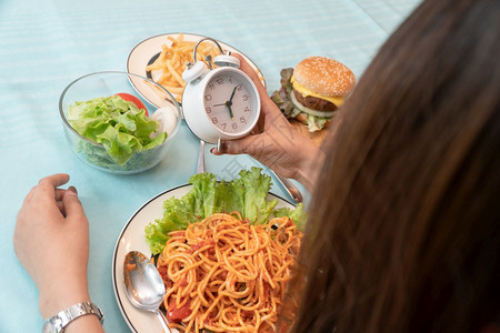 年轻女持时钟准备吃汉堡包薯条和意大利面的早餐计划狂食饮紊乱症BED和以垃圾食品不健康来放松娱乐概念快速地狂欢小吃图片