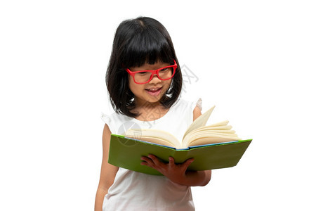 带着红眼镜的快乐亚洲学龄前小女孩拿着并阅读一本关于孤立白背景的绿皮书小学前班家庭校的童与教育概念小学家庭校在工作泰国知识图片
