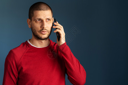 成人青年移动的穿着红色毛衣白人男子站在蓝背景墙前使用智能手机打电话聊天谈在蓝色背景墙前面用智能手机进行电话交谈图片