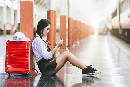 旅行的怀孕亚洲妇在火车站用红色手提箱看Smartphone车步行铁路等待图片