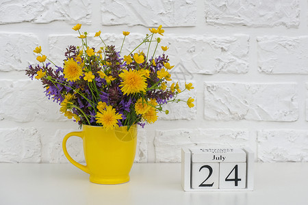 木制的明亮假期木制立方体日历7月24日和黄色杯子上面有鲜艳的花朵靠在白色砖墙上图片