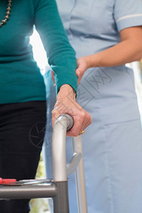八十年代老化高级女在与背景护理工作者一起行走的框架中手持重点图片