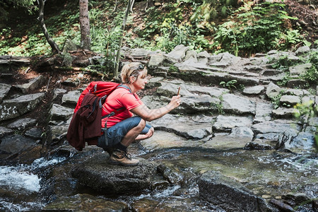 流浪癖假期积极的在夏季旅行中使用智能手机相在山上岩站立于拍摄度假女人的照片和背着包拍照的景观片图片