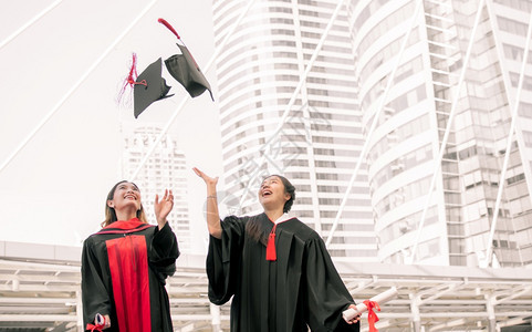 两名身穿制服的亚洲女学生在毕业后微笑着快乐和扔起帽子并带着笑容和欢的喜悦投掷庆典图片