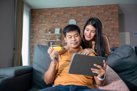 网上买亚洲情侣用笔记本电脑信卡付账在家沙发上冲网的幸福夫妇买方丈坐着图片