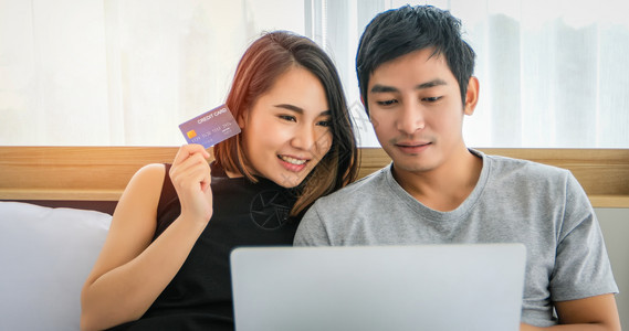 亚洲夫妇在线购物用笔记本电脑信卡付款在家床上冲网时快乐的一对夫妇银行业商购买图片