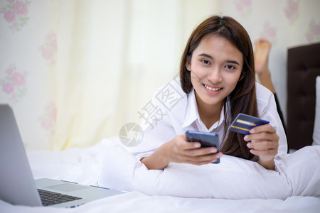 商业购买亚洲人妇女在线购物手机用信卡付账以及在家床上冲网的幸福妇女在床上买网图片